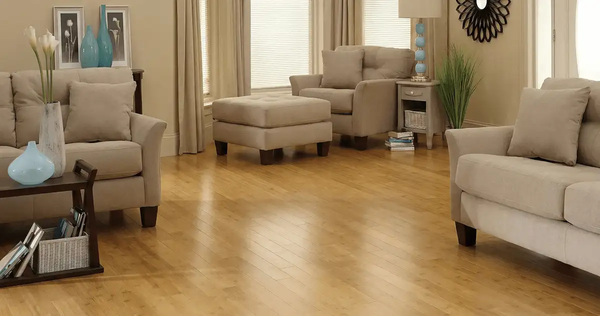 A living room with professionally installed bamboo flooring, beige sofas, a matching ottoman, a side table with a blue lamp, vases, framed pictures, and large windows with blinds and curtains in Springfield, MO